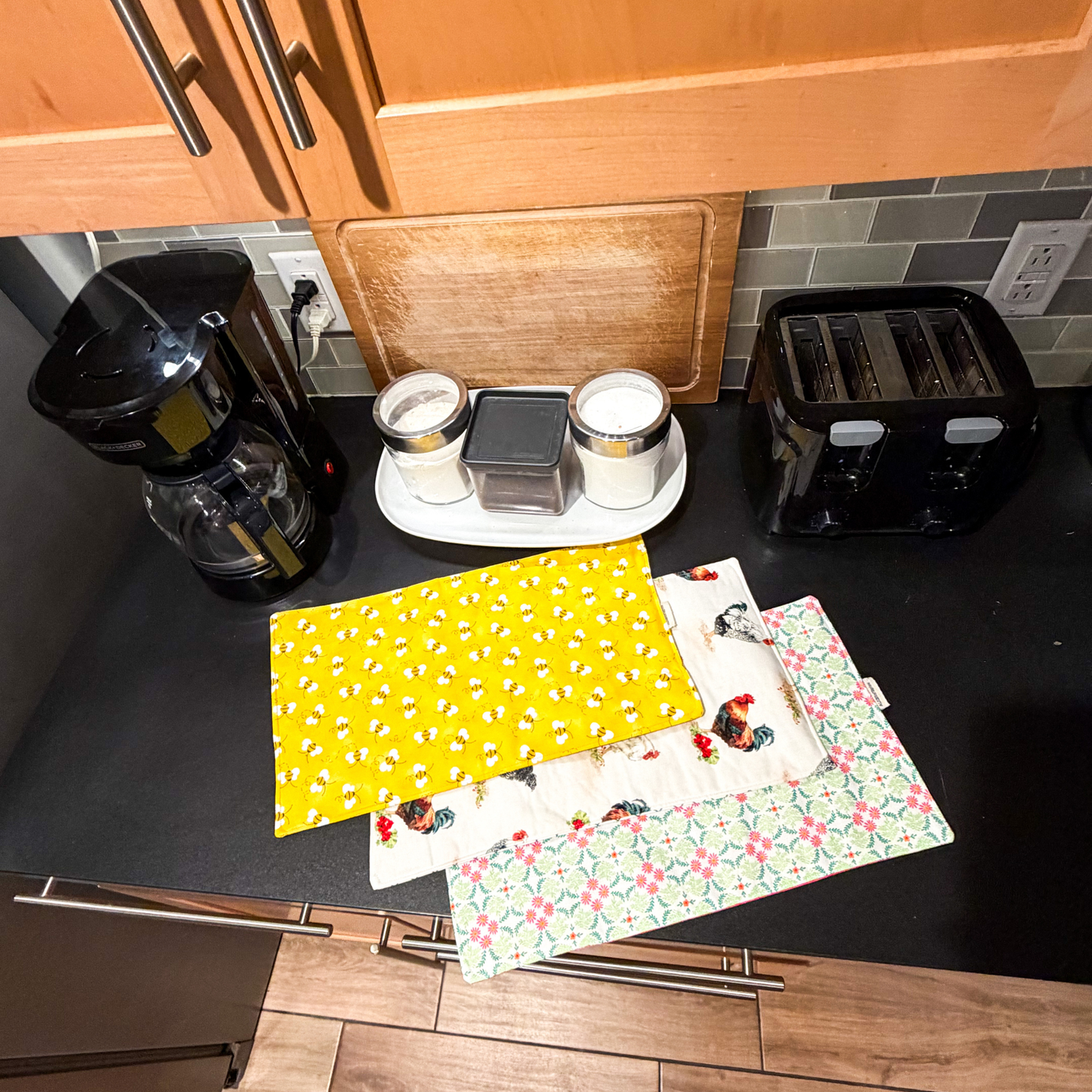 Three fabric sliding mats with different patterns are stacked on a black countertop in a kitchen setting, with a coffee maker, toaster, and cutting board visible in the background.
