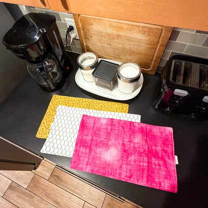 Three fabric sliding mats in pink, yellow, and white patterned styles arranged on a black kitchen countertop next to a coffee maker and toaster.