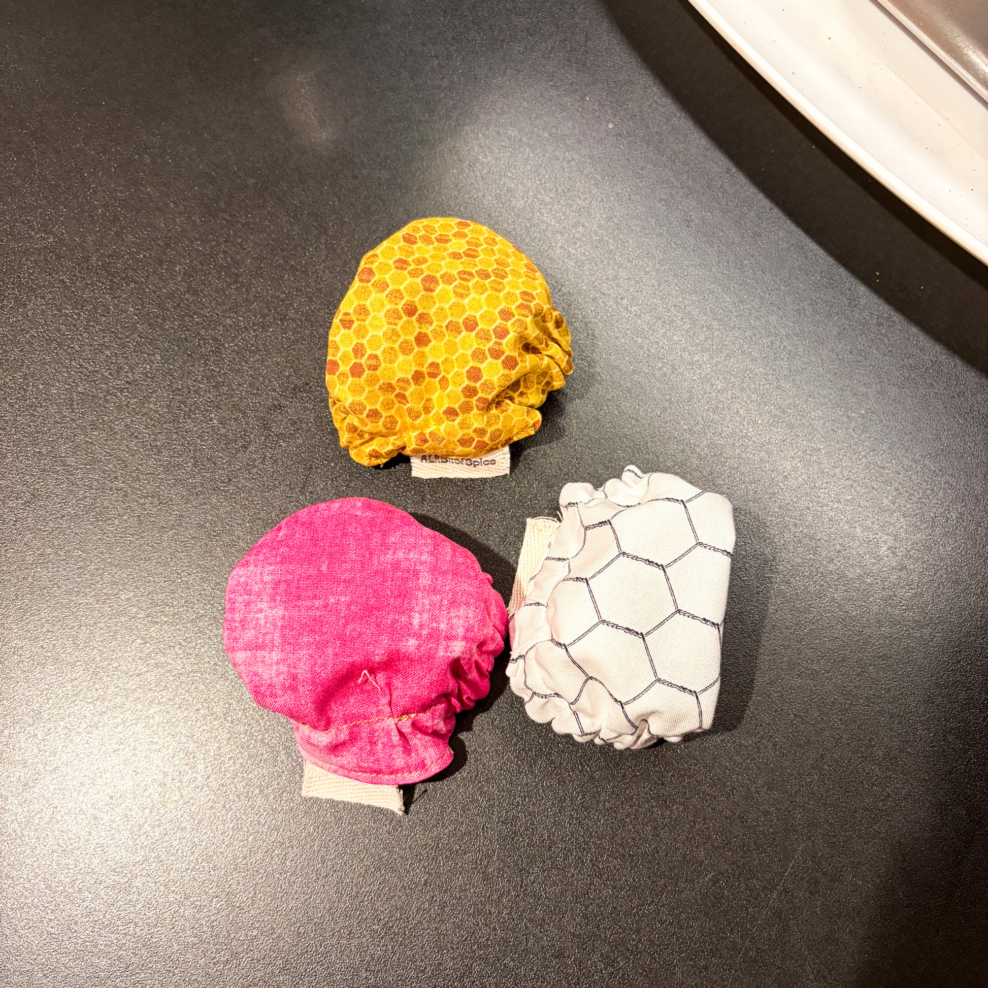 Three small fabric jar covers arranged on a dark tabletop: one yellow with a honeycomb pattern, one bright pink, and one white with a dark chicken wire pattern. Small fabric tags are visible beneath each item.