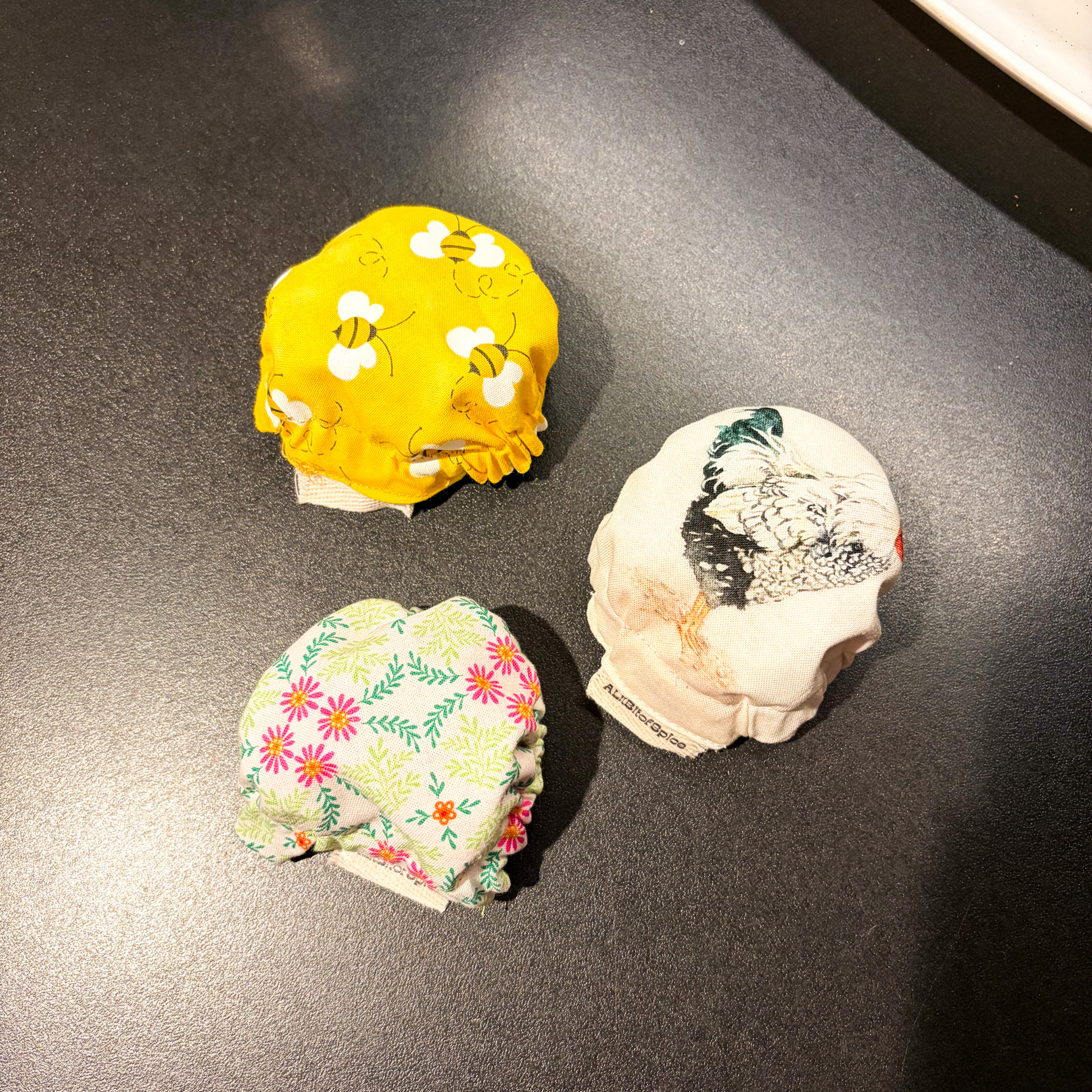 Three reusable jar covers arranged on a dark surface: one bright yellow with white bumblebees, one cream with a black-and-white rooster print, and one cream with pink and green floral patterns.