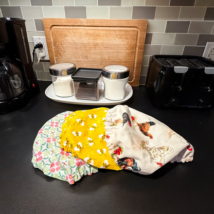 Three fabric bowl covers or burp cloths in different patterns (floral green and pink, yellow with bumblebees, and cream with rooster print) laid out on a black kitchen counter in front of a tiled backsplash.