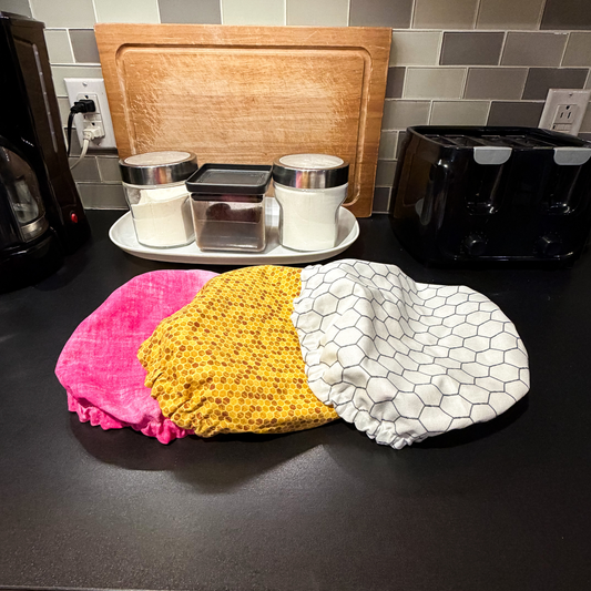Three fabric bowl covers laid out on a dark countertop: one bright pink, one yellow with a small honeycomb pattern, and one white with a black chicken wire pattern. A wooden cutting board, glass canisters, and a black toaster are visible in the kitchen background.