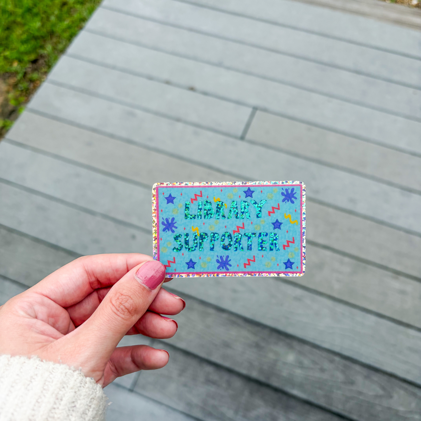 Hand holding a colorful sticker with 'Library Supporter' text on a wooden surface.
