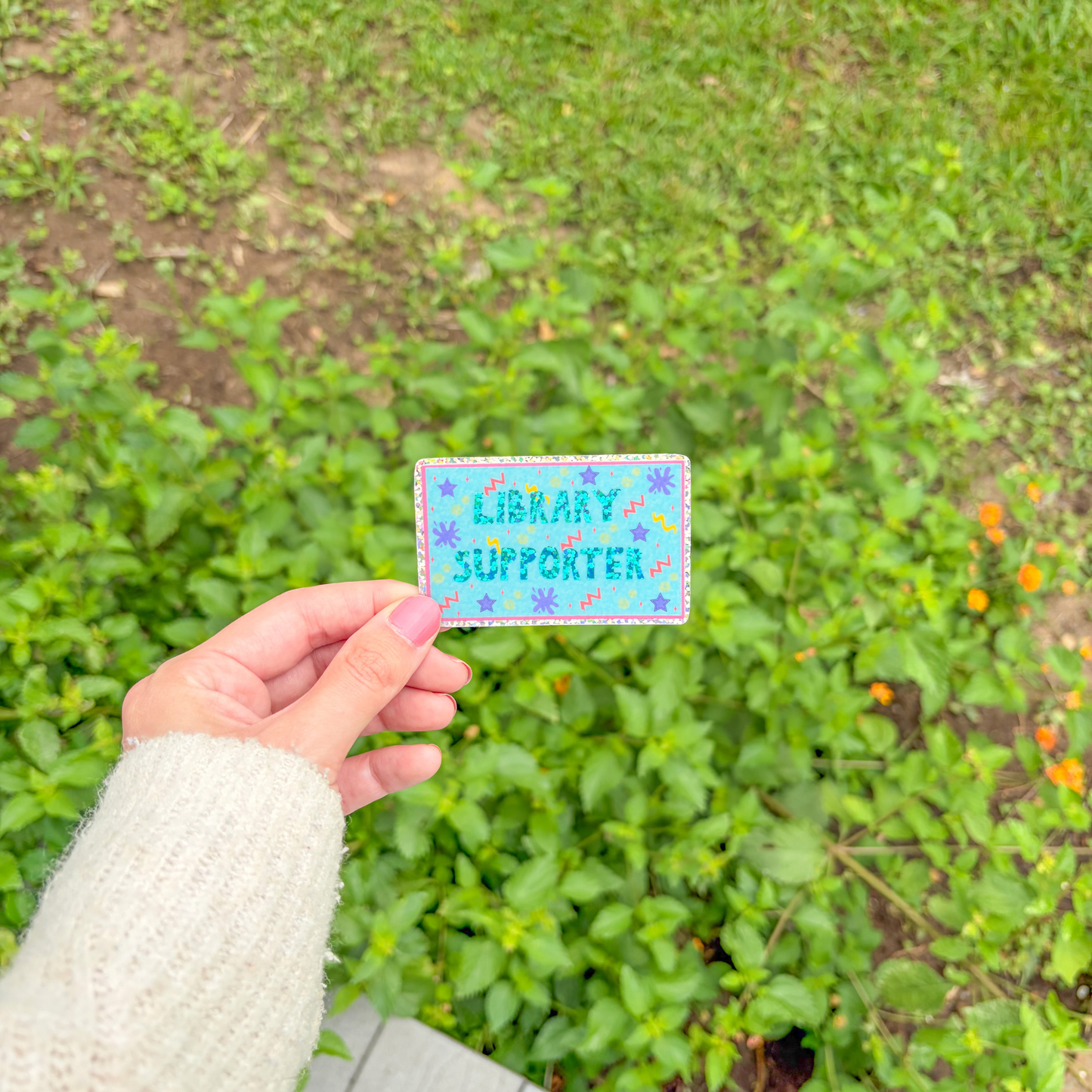 Hand holding a 'Library Supporter' patch in front of green plants