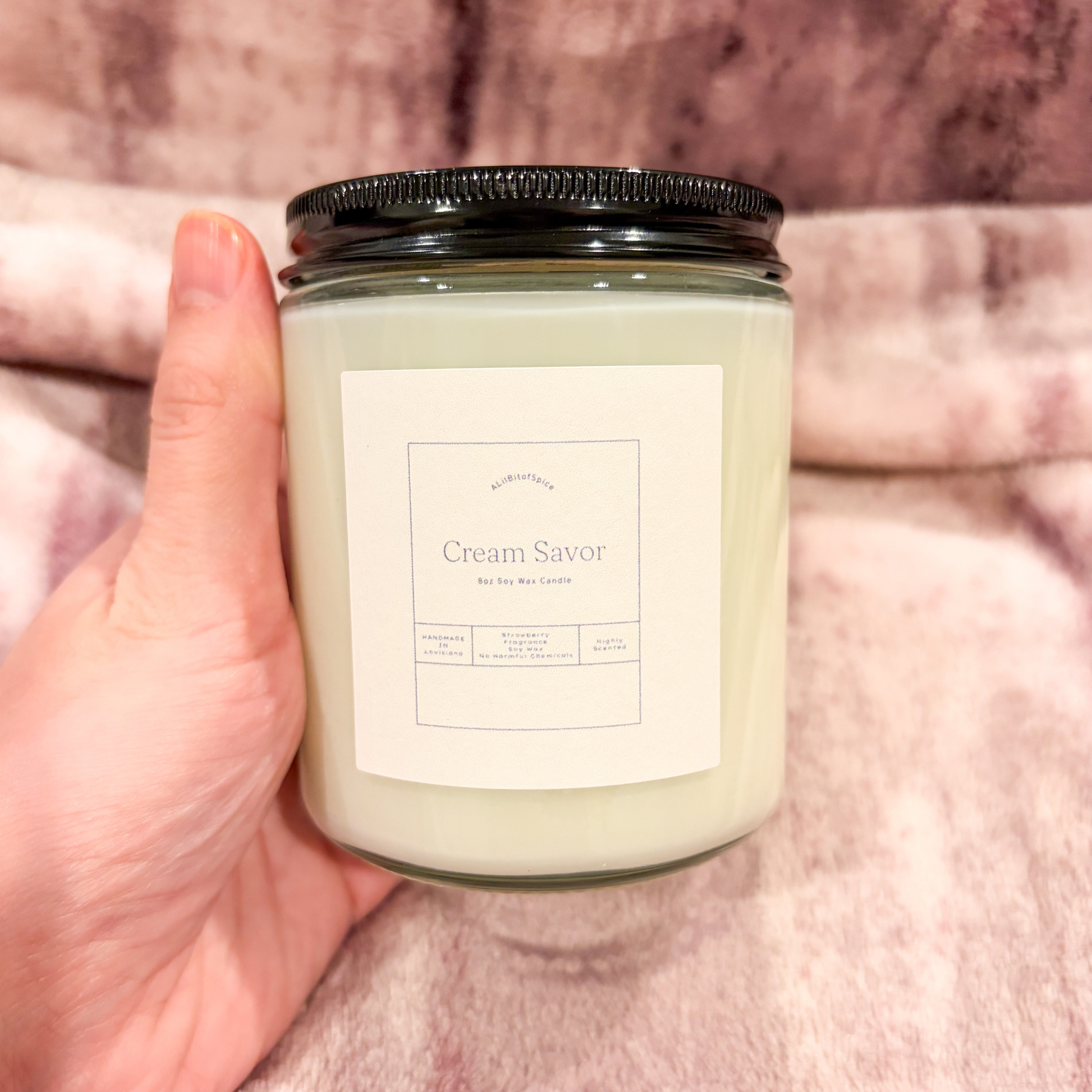 Hand holding a candle labeled 'Cream Savor' against a pink fabric background