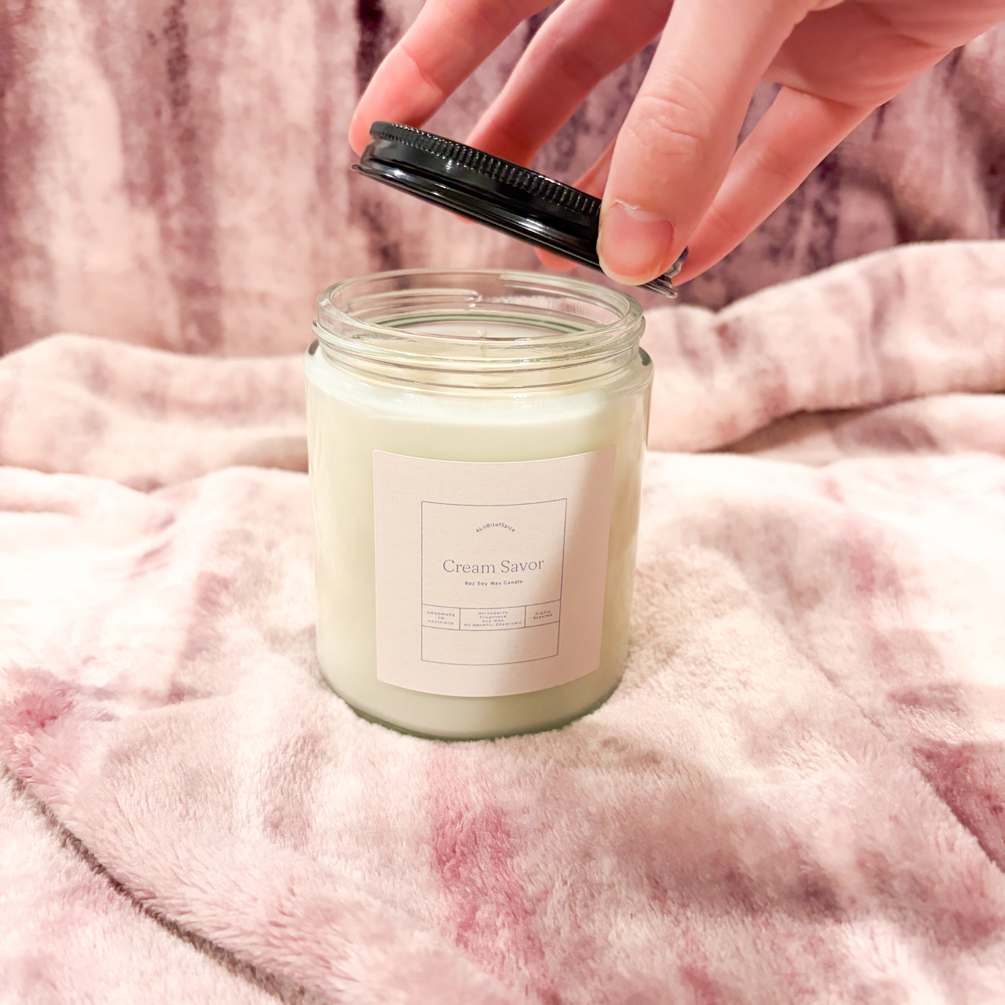 Hand opening a candle jar labeled 'Cream Savor' on a pink textured fabric background