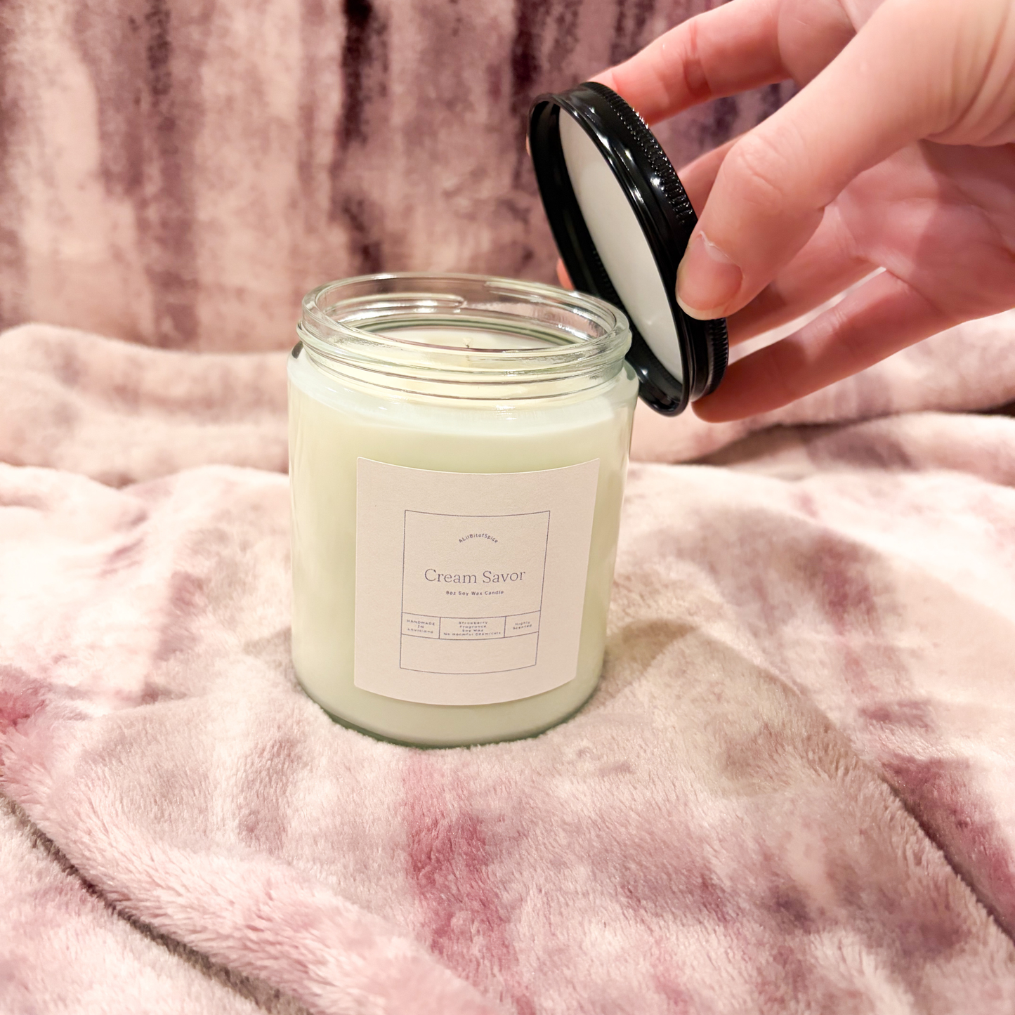 Candle jar labeled 'Cream Savor' with a hand lifting the lid on a pink textured blanket.