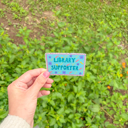 Hand holding a 'Library Supporter' patch against a green outdoor background