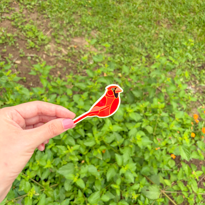 Hand holding a red cardinal sticker in front of green foliage