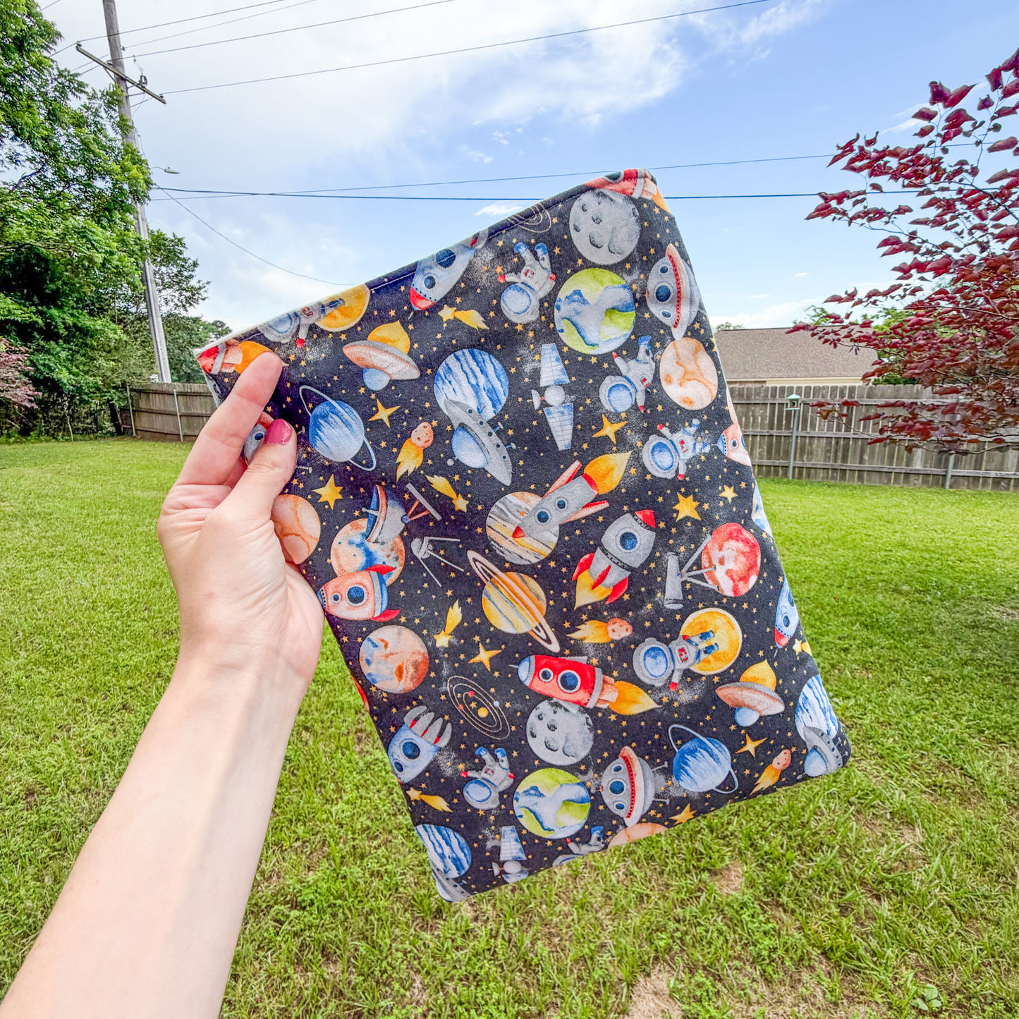 Hand holding a space-themed large book sleeve with planets, stars, and rockets against a grassy outdoor background.