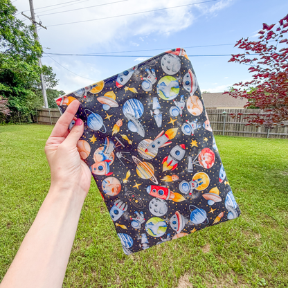 Hand holding a space-themed large book sleeve with planets, stars, and rockets against a grassy outdoor background.
