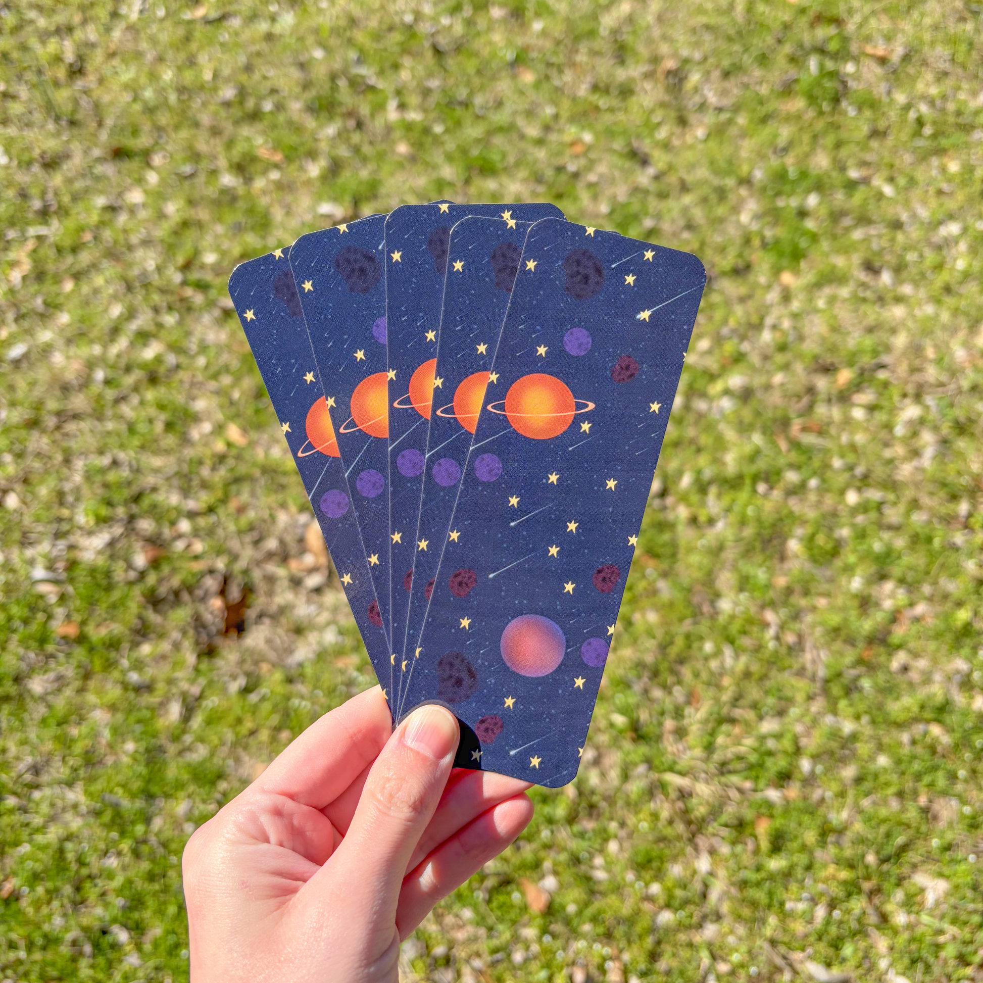 Hand holding a set of rectangular bookmarks with space-themed designs against a grassy background