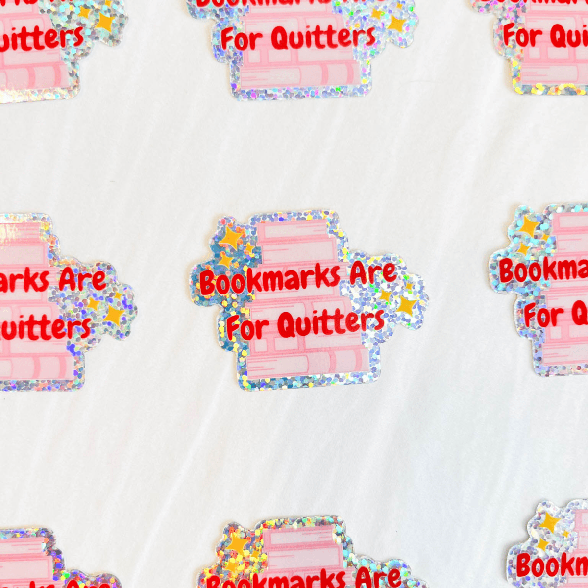 Multiple stickers laying flat with a glitter shimmer that say ‘Bookmarks Are For Quitters’ over a stack of pink books with three gold stars above and below the words on a white background.